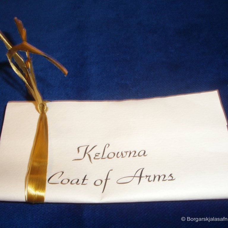 Blað með gylltu bandi utan um. Á blaðið er skrifað: Kelowna Coat of Arms ásamt texta og neðst stendur Golden Jubilee Year 1955. Kelowna er stærsta borgin í Britis Columbia´s Okanagan Valley í Kanada.